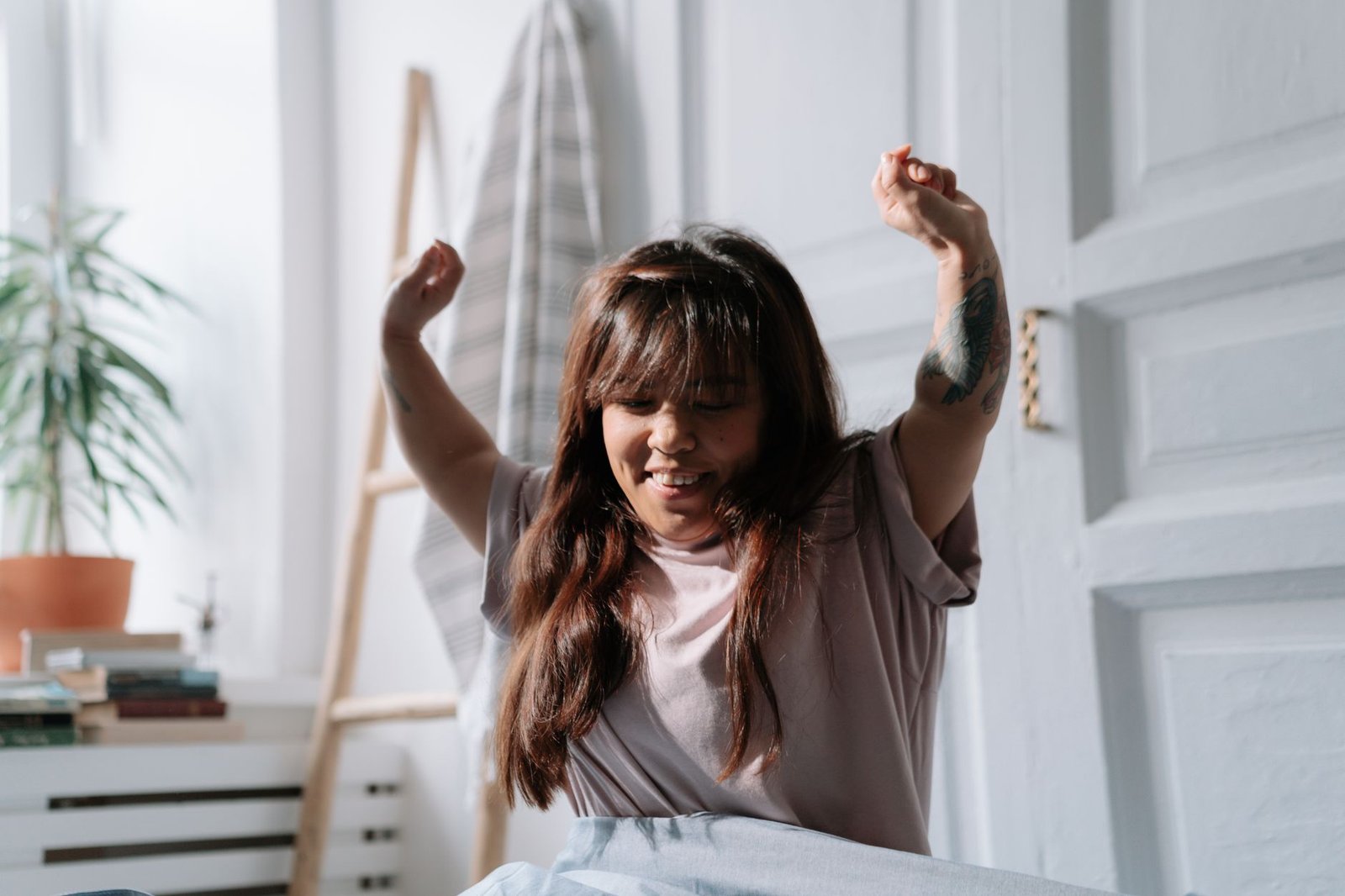woman waking up peacefully in morning sunlight stretching comfortably in a bright bedroom with white bedding and plants